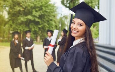 Smiling high school graduate holding a diploma in front of a group of fellow graduates in caps and gowns.