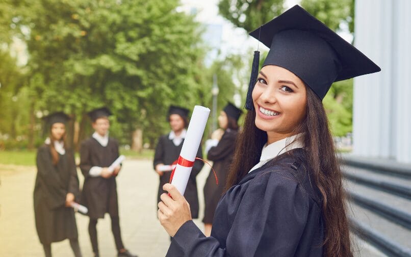 Smiling high school graduate holding a diploma in front of a group of fellow graduates in caps and gowns.